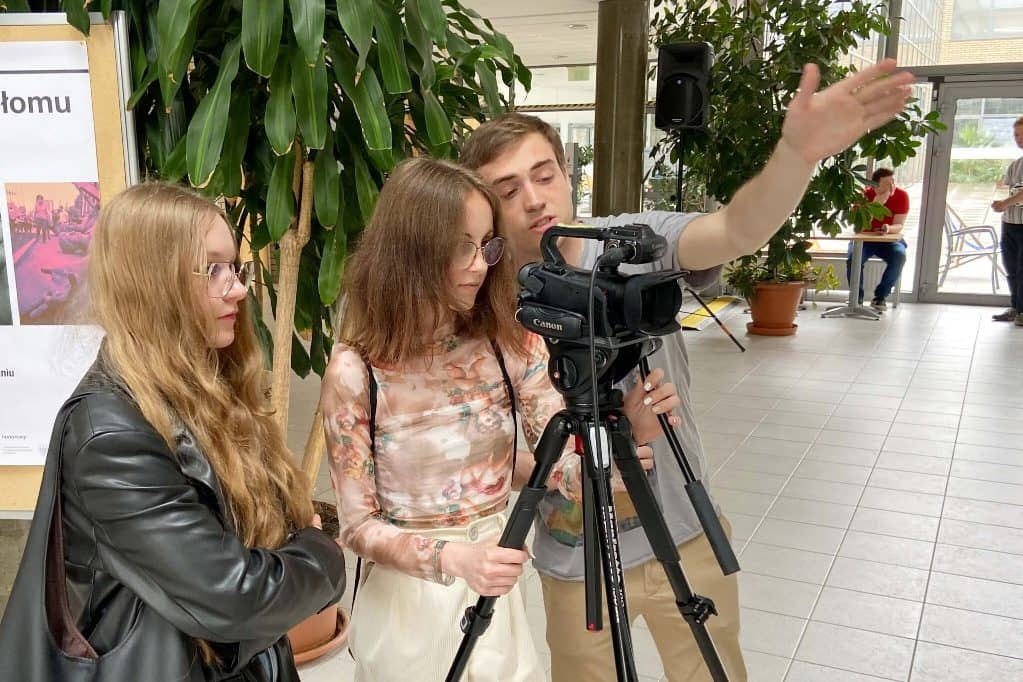 Na zdjęciu trzy osoby: instruktoró i dwie uczennice uczące się obsługiwać kamerę filmową.