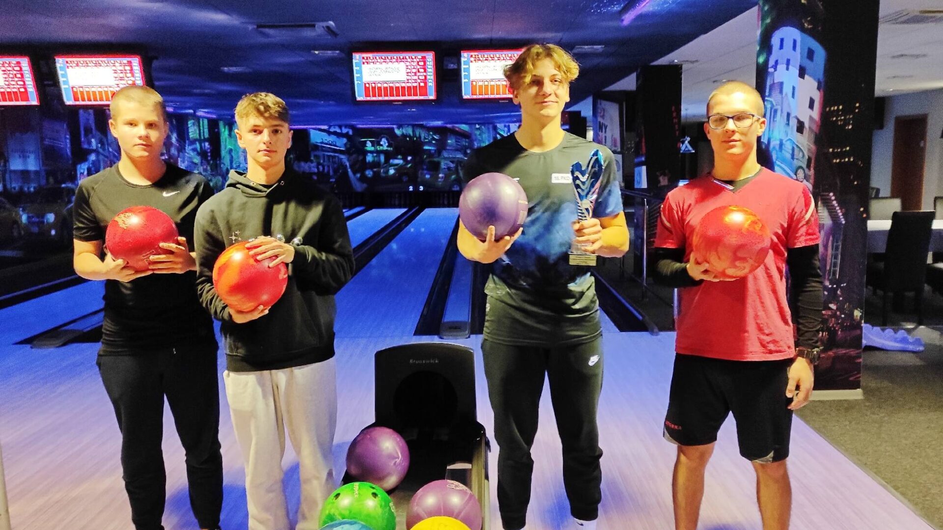 Na zdjęciu czwórka uczniów w sali bowlingowej, pozuje do zdjęcia po odebraniu nagrody za wygranie turnieju. W tle tor bowlingowy.