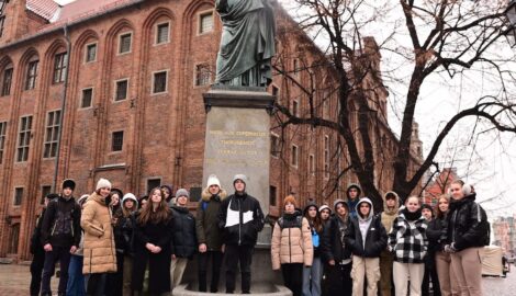 Na zdjęciu grupa uczniów w mieście Toruń. Osoby stoją na tle pomnika Mikołaja Kopernika. W tle wysoki ceglany budynek.