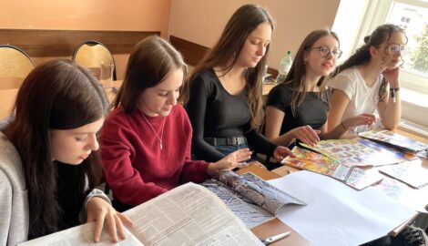 Na zdjęciu czwórka uczniów w szkolnej ławce podczas warsztatów dziennikarskich. Uczniowie przeglądają materiału z którymi będą realizować projektowe ćwiczenia.