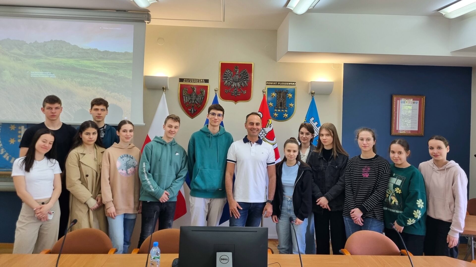 Na zdjęciu grupa uczniów wraz z pracownikiem Starostwa w Pleszewie. Osoby stoją na tle herbu powiatu.