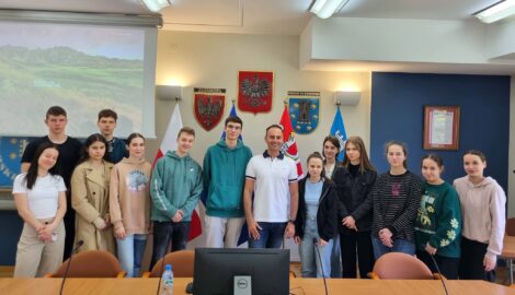 Na zdjęciu grupa uczniów wraz z pracownikiem Starostwa w Pleszewie. Osoby stoją na tle herbu powiatu.