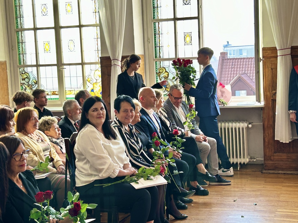 Na zdjęciu nauczyciele i zaproszeni goście siedzący w szkolnej auli. Osoby na zdjęciu trzymają kwiaty. W tle uczniowie wręczający kwiaty.