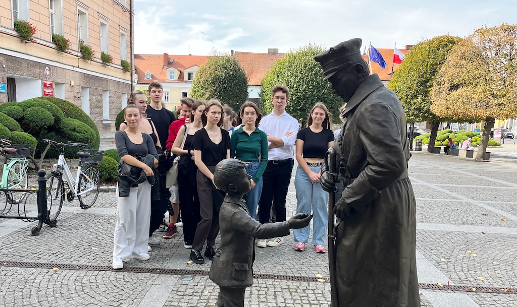 Na zdjęciu grupa uczniów na rynku w Pleszewie. Uczniowie ogladają pomnik żołnierza. Uczniowie uczestniczą w lekcji historii, w plenerze.