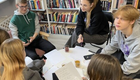Na zdjęciu uczniowie podczas poetyckich warsztatów. Uczestnicy siedzą przy stoliku i pracują nad wierszami. W tle pólki z ksiażkami.