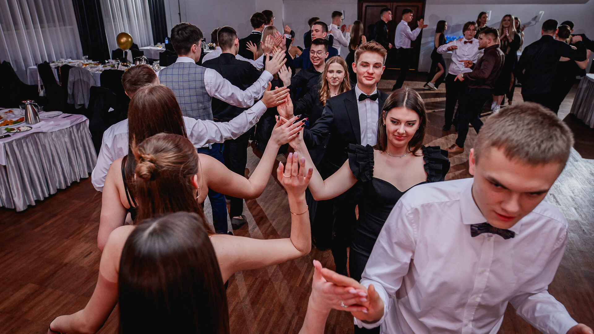 Na zdjęciu uczniowie i nauczyciele na parkiecie sali balowej, tańczą poloneza. W tle stoliki dla gości.