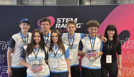Grupa uczniów reprezentujących szkołę w konkursie Steam Racing. Uczniowie pozują do zdjęcia wraz opiekunką na tle banner z logo konkursu.