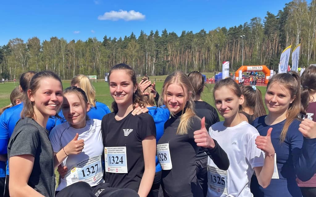 Na zdjęciu szkolna drużyna biegaczek. Szóstka dziewcząt pozuje do zdjęcia w sportowych sporach na tle lasu, gdzie rozgrywano zawody w biegach przełajowych.