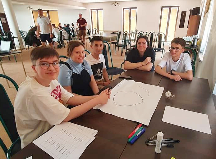 PIątka uczestników projektu Erasmus siedzi przy stole i realizuje zadanie projektowe.