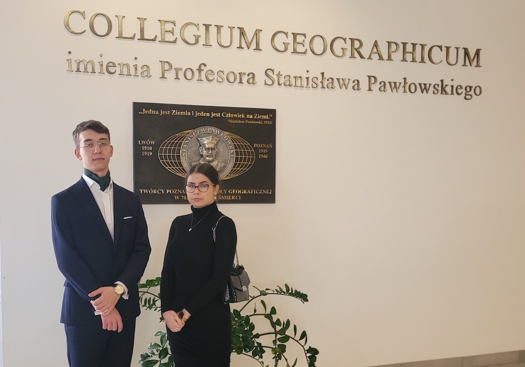 Na zdjęciu dwoje uczniów, uczestników Olimpiady Geograficznej. Uczniowie pozują do zdjęcia w Collegium Geographicum na UAMie w Poznaniu, na tle pamiątkowej tablicy.