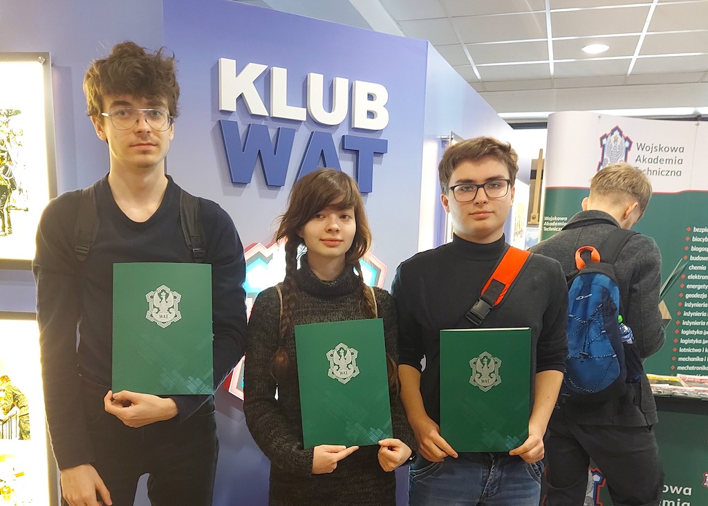 Na zdjęciu trójka uczniów z dyplomami uzyskanymi w matematycznym konkursie. W tle banner z napisem Klub Wat.