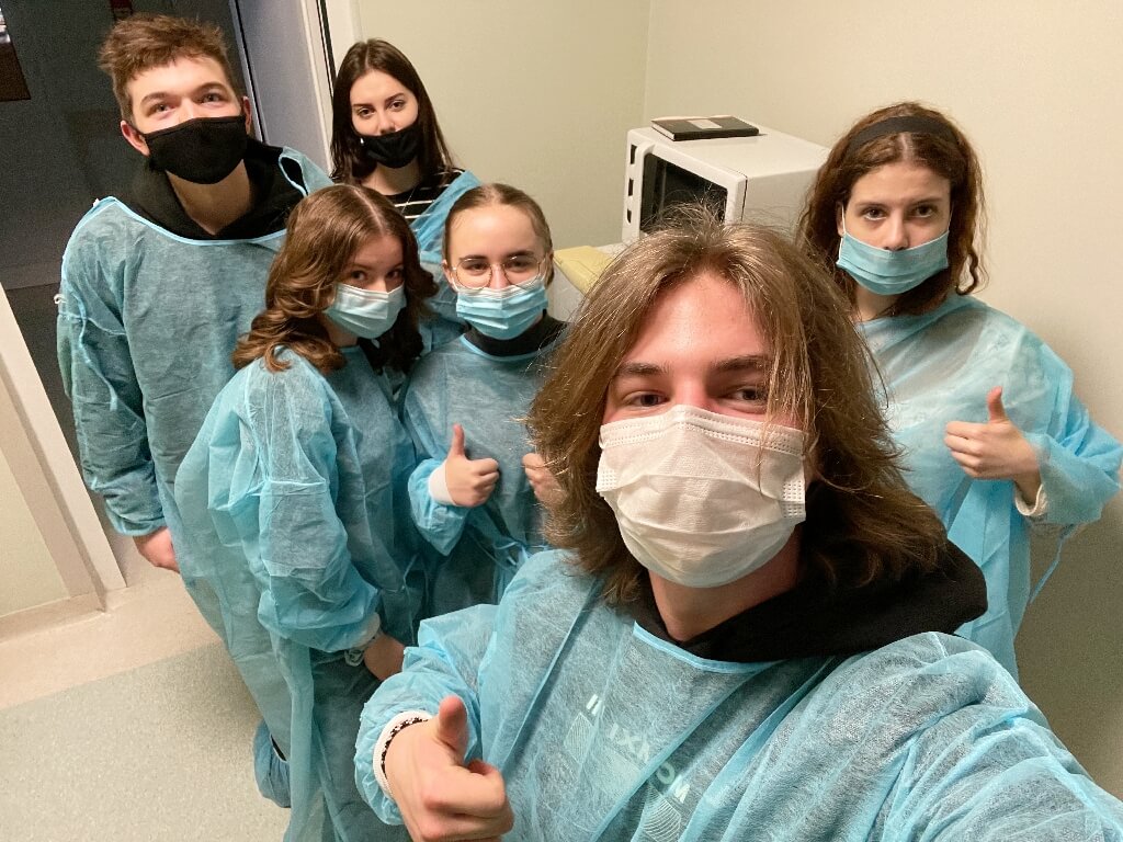 Na zdjęciu grupa uczniów w kitlach i maseczkach, podczas warsztatów w lokalnym szpitalu. Uczniowie robią selfie.
