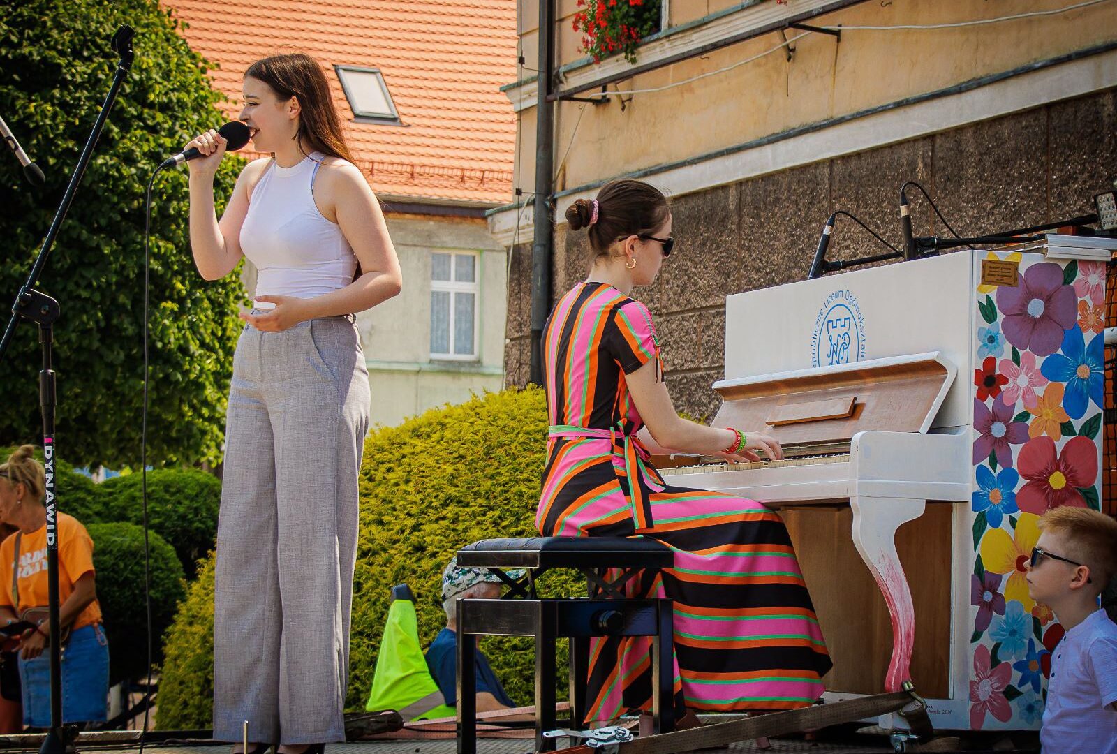 Na zdjęciu uczennice na zaimprowizowanej scenie, przed ratuszem miejskim w Pleszewie. Uczennice wykonują utwór muzyczny w ramach akcji: wędrujące pianino.