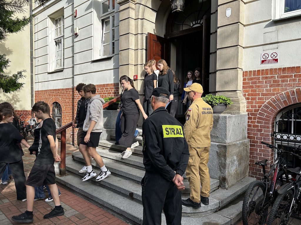 Na zdjęciu uczniowie i nauczyciele wychodzący z budynku szkoły podczas ćwiczeń w ewakuacji budynku. Na pierwszym planie uczestniczący w akcji strażacy.