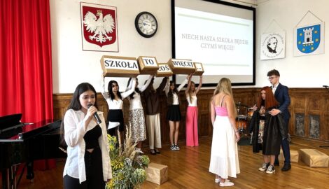 Na zdjęciu grupa kliku uczniów na scenie szkolnej auli. Uczniowie występują podczas przedstawienia otwierającego rok szkolny. Jedna uczennica z mikrofonem wykonuje piosenkę. W tle, na ścianie widoczny slajd prezentacji z hasłem: Niech nasza szkoła będzie czymś więcej.