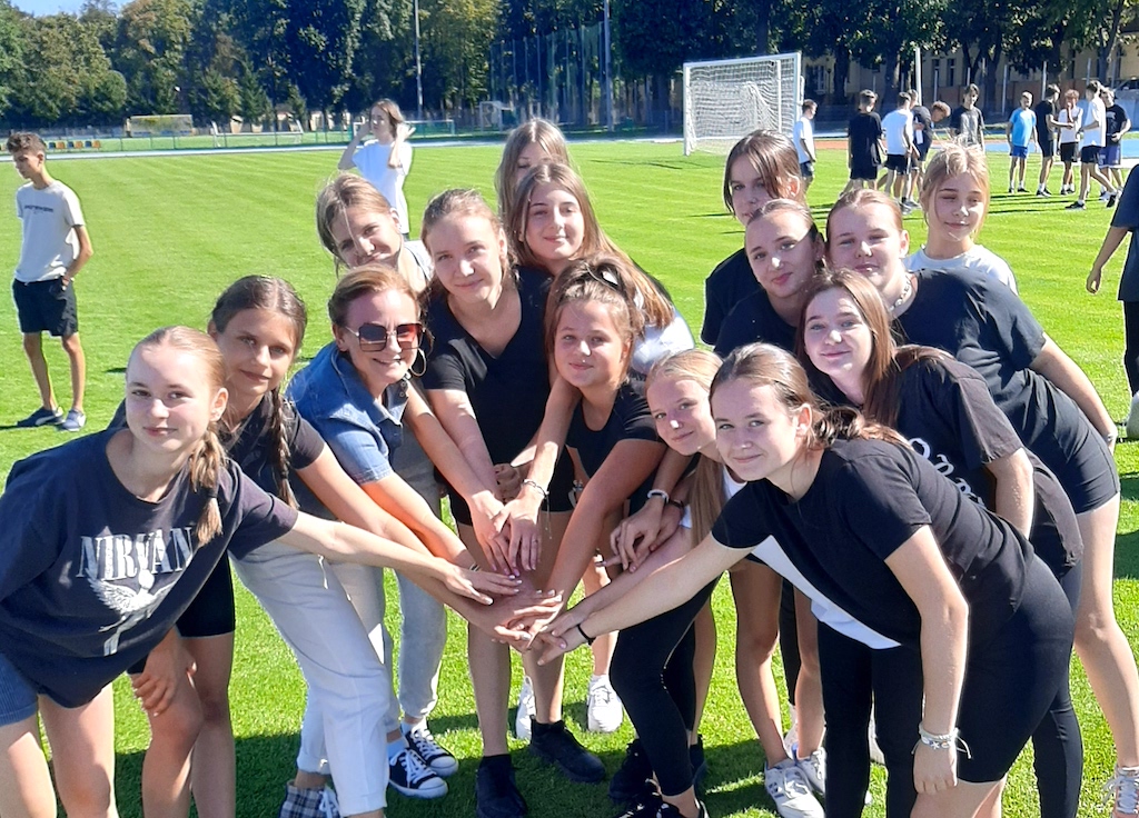 Na zdjęciu grupa uczennic na murawie boiska piłkarskiego, podczas szkolnej olimpiady. W tle uczniowie przygotowujący się do konkurencji sportowej.